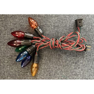 Puede incluir: Una cadena de bombillas incandescentes multicolores en naranja, rojo, rojo oscuro, verde, azul y amarillo. Las bombillas están unidas a un cable eléctrico trenzado verde y rojo con un enchufe negro y un conector de extremo a extremo negro.