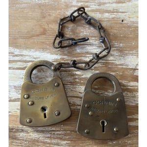 May include: Two antique brass padlocks with a dark, weathered patina. Each padlock is engraved with the text CORBIN SECURE LEVER and features a keyhole. One padlock is attached to a rusted, dark metal chain with several interconnected links.