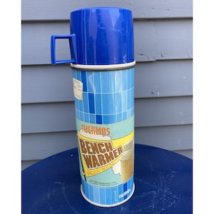 Może przedstawiać: Termos vintage Thermos Bench Warmer z niebieską plastikową zakrętką w formie kubka z uchwytem oraz cylindrycznym korpusem ozdobionym niebieskim wzorem w kratkę. Etykieta zawiera napis Thermos Bench Warmer Vacuum Bottle w kolorze pomarańczowym i brązowym.