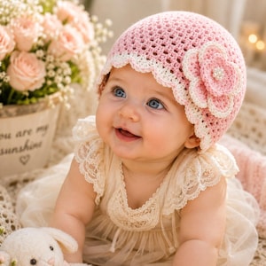 May include: A light pink crochet baby hat featuring a scalloped white trim and a large, layered pink and white crochet flower accent with a central cluster of small white beads, worn by a baby in a cream-colored lace-trimmed dress.
