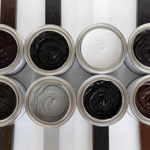 May include: Eight open circular metal paint tins arranged in two rows, each containing a different coloured paint with a visible swirl pattern. The colours include shades of black, white, grey, and brown. Four paint brushes are placed at the corners.