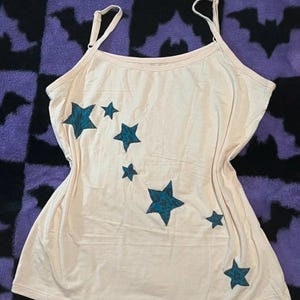 May include: A light cream-coloured camisole tank top featuring thin adjustable shoulder straps and a scoop neckline. The front of the garment is decorated with a series of six dark teal stars of varying sizes arranged in a curved, diagonal pattern.