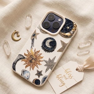 May include: A white smartphone case decorated with intricate beadwork featuring a golden sun, various stars, and crescent moons in shades of blue, gold, and silver. The case includes a tag with the text bliss and bling printed in gold script.