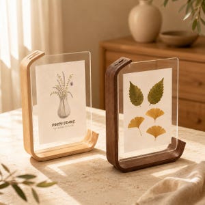 Puede incluir: Dos marcos de fotos flotantes con bases de madera clara y oscura. El marco izquierdo contiene una impresión de un jarrón con flores y el texto 5 x 7 in (13 x 18 cm). El marco derecho muestra cuatro hojas prensadas. Paneles de acrílico.