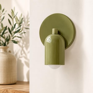 Op de afbeelding: Een wandlamp met een ronde, olijfgroene achterplaat en een bijpassende cilindrische kap met een afgeronde bovenkant. De armatuur bevat een kleine, witte, halfronde lamp die zichtbaar is bij de onderste opening van de groene metalen behuizing.
