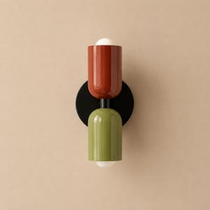 May include: A wall-mounted light fitting featuring a circular black base and two cylindrical shades. The top shade is a glossy terracotta red, and the bottom shade is a glossy olive green. Each shade contains a small, rounded white bulb.