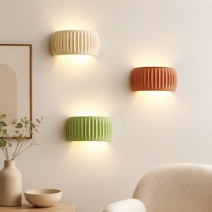 Pode incluir: Três arandelas semicirculares com texturas caneladas verticais, iluminadas de cima e de baixo. As luminárias são nas cores creme, terracota e verde sálvia, montadas em uma parede, projetando um brilho quente na superfície.