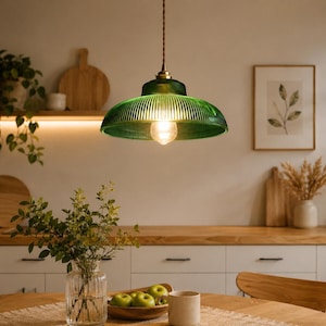 Könnte beinhalten: Eine Pendelleuchte mit einem breiten, gerippten, smaragdgrünen Glasschirm und einer messingfarbenen Metallfassung. Die Lampe hängt an einem gedrehten braunen Kabel und enthält eine leuchtende Glühbirne, die das Glas erhellt.