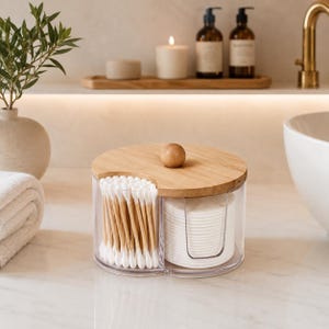 Op de afbeelding: Een doorzichtige, ronde opbergcontainer van acryl met een verdeelde binnenkant en een rond houten deksel. De ene kant bevat een verticale stapel witte wattenstaafjes met houten steeltjes, de andere kant bevat ronde witte wattenschijfjes.