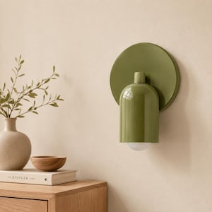 Puede incluir: Un aplique de pared con una placa trasera circular de color verde oliva y una pantalla cilíndrica a juego con la parte superior redondeada. La lámpara incluye una pequeña bombilla blanca semiesférica visible en la base de la carcasa.