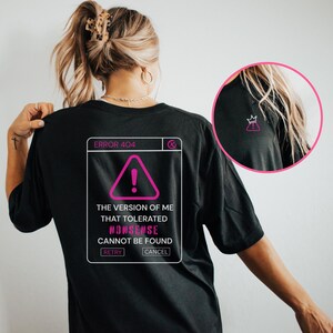 May include: A black t-shirt featuring a white and pink graphic on the back that resembles a computer error window. The text reads ERROR 404, THE VERSION OF ME THAT TOLERATED NONSENSE CANNOT BE FOUND, RETRY, and CANCEL. A small crown icon is visible.