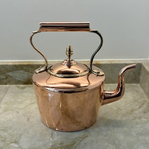 May include: A polished copper tea kettle featuring a rounded body, a curved spout, and a hinged handle. The lid is topped with a small, decorative finial. The metal surface reflects light, showcasing a warm, metallic orange-brown hue throughout.