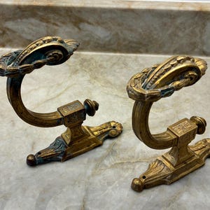 May include: Two matching brass curtain tiebacks featuring a curved, C-shaped arm with an ornate, laurel-leaf inspired top detail. The metal has a weathered, aged finish with visible dark patina in the recessed areas and a rectangular base.