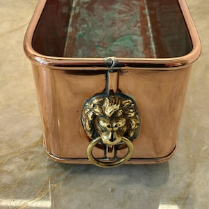 May include: A rectangular copper planter with rounded corners and a polished finish. The exterior features a prominent brass lion head ornament centred on the side, which holds a circular brass ring handle. The interior shows signs of oxidation.