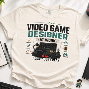 May include: A cream-colored t-shirt featuring a graphic print that reads "VIDEO GAME DESIGNER AT WORK" and "I DON'T JUST PLAY." The design includes icons for imagine, design, code, create, test, and improve, surrounding a computer desk illustration.