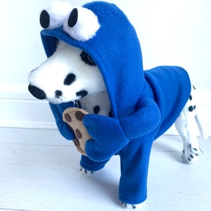 May include: A Dalmatian dog wearing a blue hooded costume with large white eyes and holding a chocolate chip cookie. The costume is designed to look like a cookie monster.