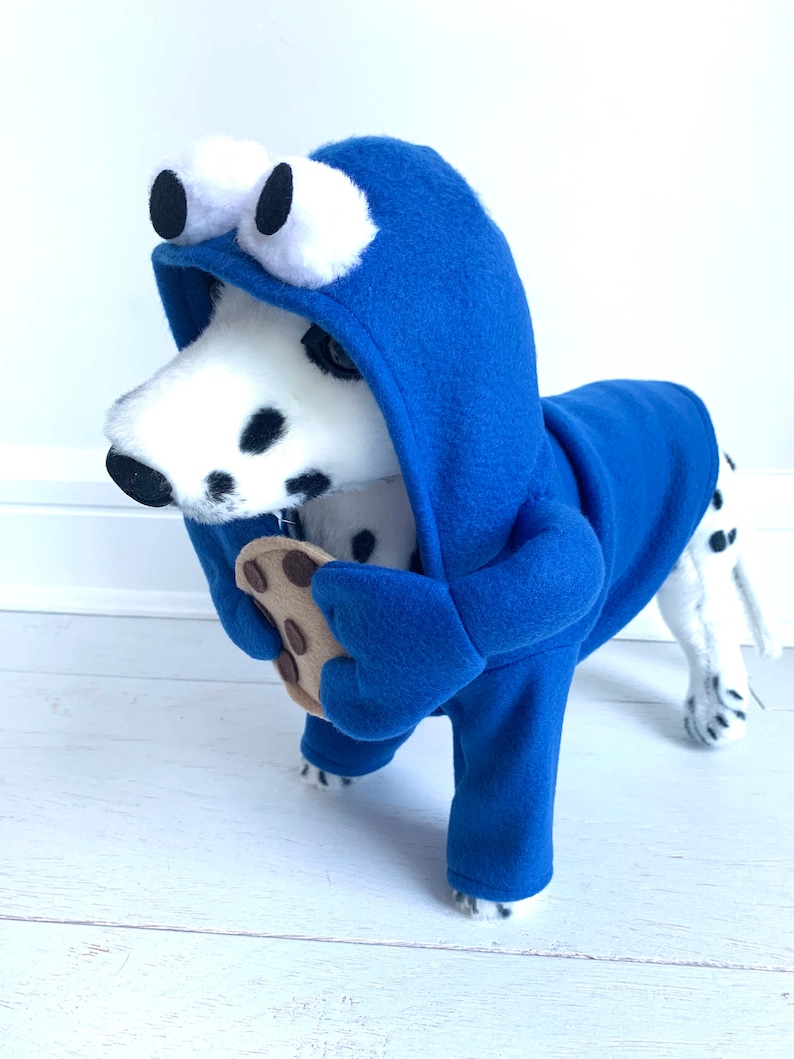 May include: A Dalmatian dog wearing a blue hooded costume with large white eyes and holding a chocolate chip cookie. The costume is designed to look like a cookie monster.