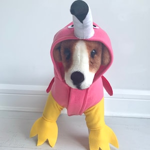 May include: A dog wearing a pink flamingo costume. The costume features a pink hood with a white and black beak, pink wings, and yellow legs with webbed feet. The dog has brown and white fur.