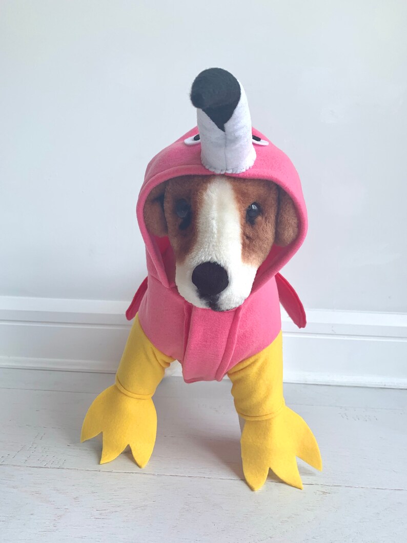 May include: A dog wearing a pink flamingo costume. The costume features a pink hood with a white and black beak, pink wings, and yellow legs with webbed feet. The dog has brown and white fur.