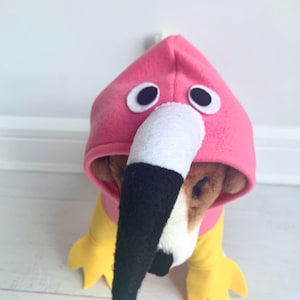 May include: A dog wearing a pink flamingo costume. The costume features a pink hood with large white-rimmed eyes and a white and black beak. The dog's body is covered in a yellow and pink outfit.