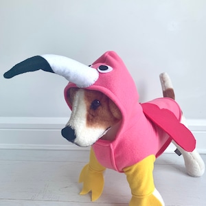 May include: A dog wearing a pink flamingo costume. The costume features a pink hood with a white and black beak, pink wings, and yellow feet. The dog is standing on a white surface.