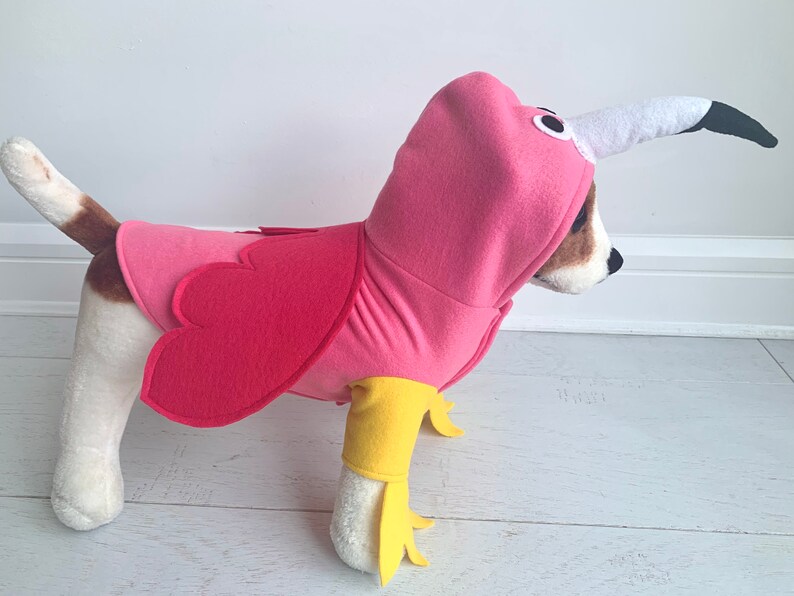 May include: A dog wearing a pink flamingo costume. The costume includes a pink hooded top with a white and black beak, and pink wings. The dog's legs are covered with yellow fabric, resembling flamingo legs.