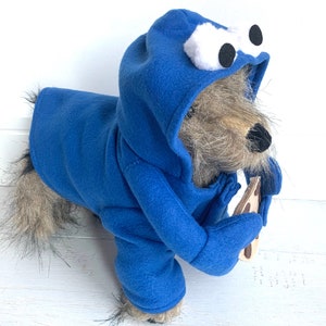 May include: A blue dog costume with a cookie monster design. The costume has a hood with white eyes and a brown cookie in the paw.