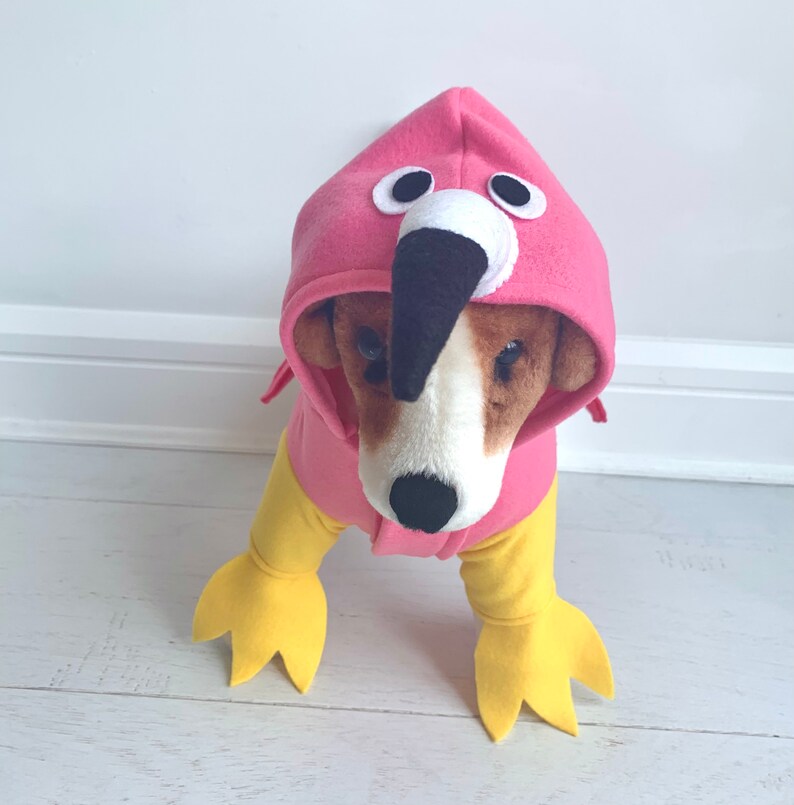 May include: A dog wearing a pink flamingo costume with a hood, beak, and eyes. The costume has yellow legs and feet. The dog is standing on a white wooden floor.