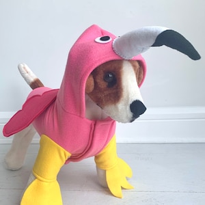 May include: A dog wearing a pink flamingo costume with a hood, beak, and wings. The costume has yellow feet and a zipper down the front. The dog is standing on a white surface.