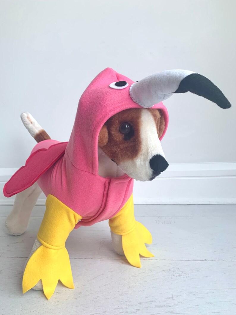 May include: A dog wearing a pink flamingo costume with a hood, beak, and wings. The costume has yellow feet and a zipper down the front. The dog is standing on a white surface.