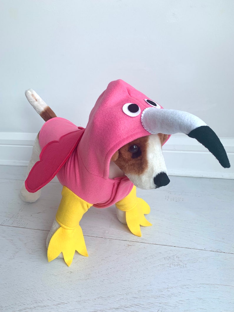 May include: A dog wearing a pink flamingo costume with a hood, wings, and yellow feet. The costume has a white beak with a black tip and large black and white eyes. The dog is standing on a white wooden floor.
