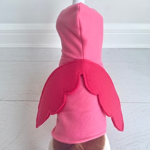 May include: A pink flamingo dog costume. The costume features a pink hooded top with a white and black flamingo head and a pair of red wings on the back. The dog's tail is visible.