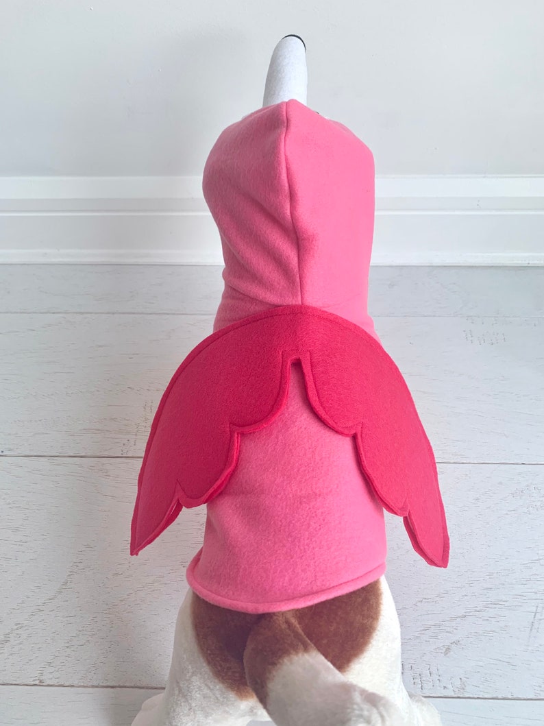 May include: A pink flamingo dog costume. The costume features a pink hooded top with a white and black flamingo head and a pair of red wings on the back. The dog's tail is visible.