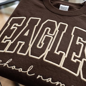 May include: A dark brown crewneck sweatshirt featuring the word EAGLES in large, cream-colored, embroidered block letters. Below the main text, the phrase school name is embroidered in a smaller, cream-colored, cursive script font.