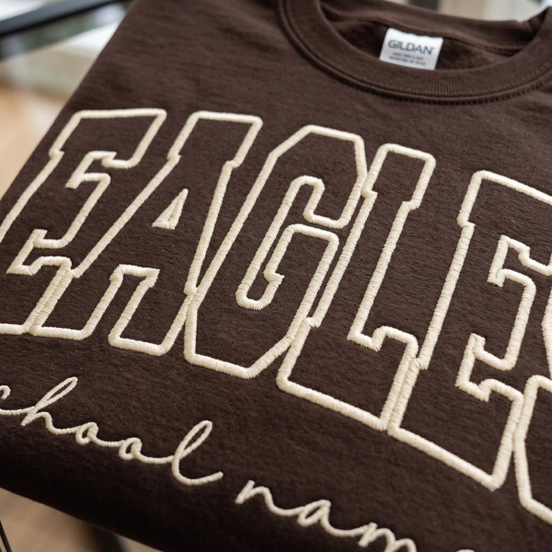 May include: A dark brown crewneck sweatshirt featuring the word EAGLES in large, cream-colored, embroidered block letters. Below the main text, the phrase school name is embroidered in a smaller, cream-colored, cursive script font.