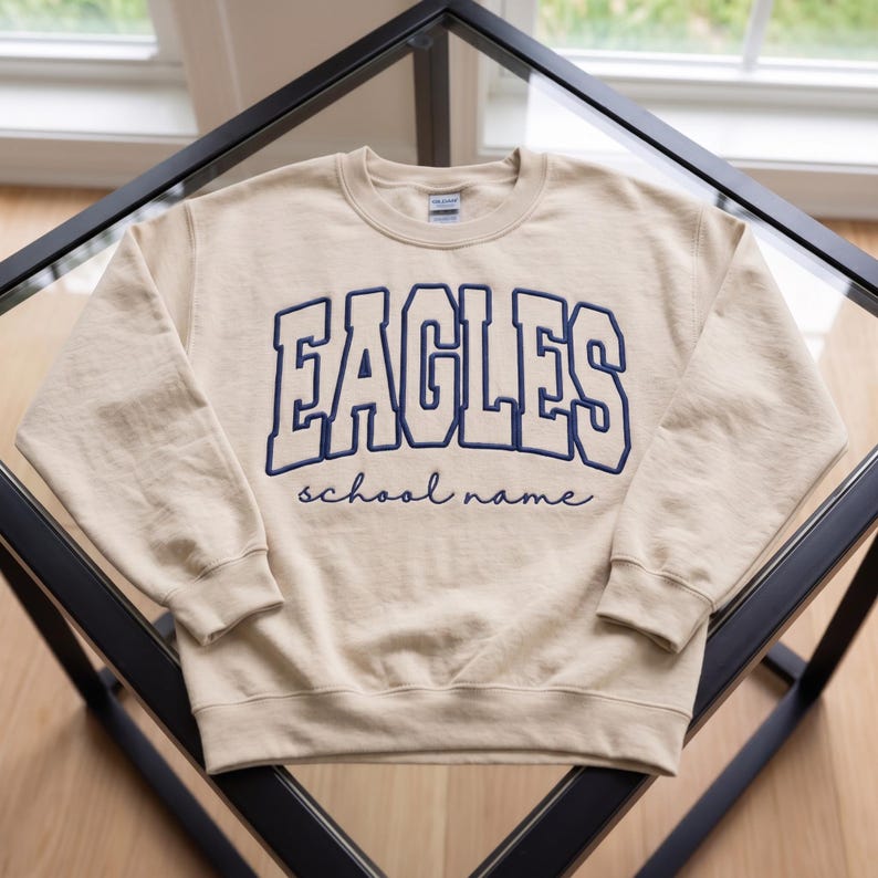 May include: A beige crewneck sweatshirt featuring the word EAGLES in large, dark blue block letters across the chest, with the words school name written in a smaller, dark blue cursive script directly underneath the block text.