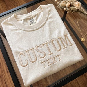 Peut inclure: Un t-shirt à col rond blanc cassé avec le mot CUSTOM brodé en grandes lettres beiges de style bloc, et le mot TEXT brodé en dessous en plus petites lettres. L'étiquette intérieure du col affiche la marque Comfort Colors.
