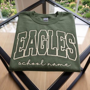 May include: An olive green crewneck sweatshirt featuring the word EAGLES in large, cream-colored block letters with a dark outline. Below the main text, the phrase school name is embroidered in a white cursive script font on the chest.