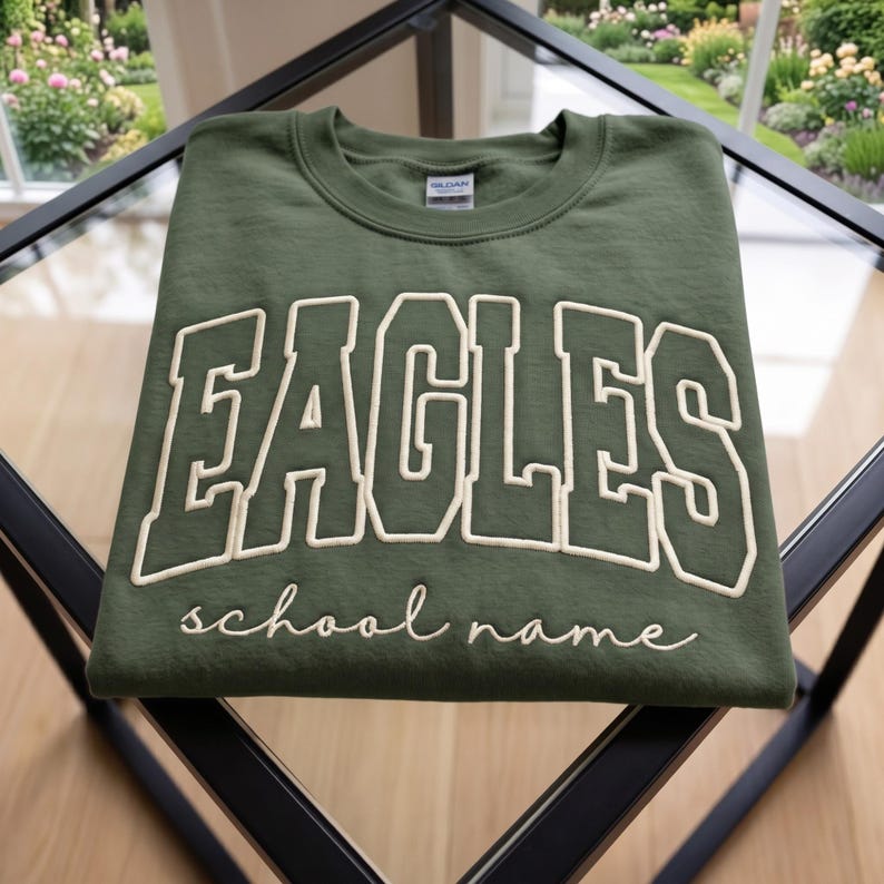May include: An olive green crewneck sweatshirt featuring the word EAGLES in large, cream-colored block letters with a dark outline. Below the main text, the phrase school name is embroidered in a white cursive script font on the chest.