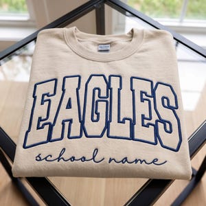 May include: A beige crewneck sweatshirt featuring the word EAGLES in large, dark blue block letters with a thick outline. Below the word, the phrase school name is embroidered in a dark blue cursive script. The garment has a ribbed collar.