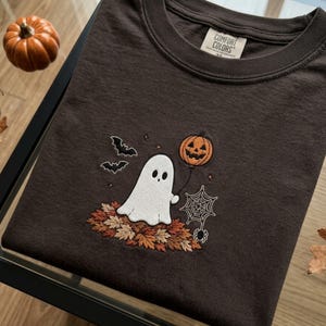 Könnte beinhalten: Ein dunkelbraunes T-Shirt mit einer Stickerei, die einen weißen Geist auf einem Haufen Herbstlaub zeigt. Der Geist hält eine Schnur, die an einem orangefarbenen Kürbisballon befestigt ist, sowie zwei schwarze Fledermäuse, ein Spinnennetz und eine Spinne.