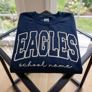May include: A navy blue crewneck sweatshirt featuring the word EAGLES in large, white, block-style embroidered lettering across the chest, with the words school name in a white cursive script font embroidered directly underneath the main text.
