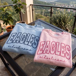 May include: A light blue sweatshirt and a light pink sweatshirt, both featuring the word EAGLES in large block letters and the text school name in cursive script. The blue sweatshirt has dark blue embroidery, while the pink one has dark pink embroidery.