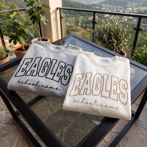 May include: Two crewneck sweatshirts, one light gray and one cream, each featuring the word EAGLES in a large collegiate font and the text school name in a cursive script. The gray sweatshirt has black embroidery, while the cream one has tan.
