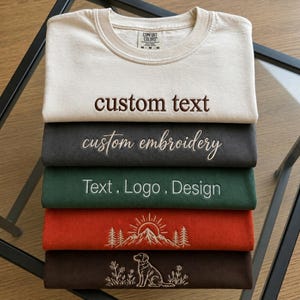Puede incluir: Una pila de cinco camisetas de cuello redondo dobladas en color crema, carbón, verde oscuro, naranja óxido y marrón oscuro. La camiseta crema tiene el texto custom text, la carbón custom embroidery, la verde Text, Logo, Design, la naranja una montaña y la marrón un perro.