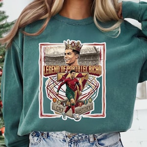 May include: A dark teal crewneck sweatshirt featuring a graphic print of a soccer player in a red and green uniform. The design includes the text "LEGEND OF PORTO | EL BICHO" and "CR7 HISTORY" with an illustration of a crowned athlete and a soccer ball.