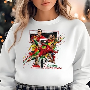 May include: A white long-sleeve sweatshirt featuring a graphic print of soccer player Cristiano Ronaldo in various action poses and portraits, accented with red, green, and yellow paint splatters and the text Cristiano Ronaldo.