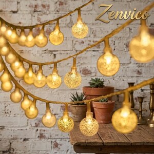 May include: A string of warm white LED globe lights with a textured, speckled finish on the glass bulbs. The lights are arranged in multiple draped strands, emitting a soft glow. The word Zenvico is printed in the upper right corner.