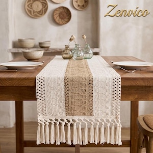 Puede incluir: Un camino de mesa rectangular con un panel central tejido en color tostado flanqueado por secciones de encaje de ganchillo color crema. El camino termina en una hilera de largos flecos color crema. Se muestra sobre una mesa de madera.