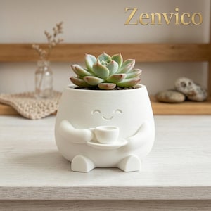 Op de afbeelding: Een witte keramische plantenpot in de vorm van een klein figuurtje met armen die een minuscuul kopje en schoteltje vasthouden. De pot heeft een lachend gezicht en bevat een groene vetplant. De tekst Zenvico is zichtbaar in de rechterbovenhoek.